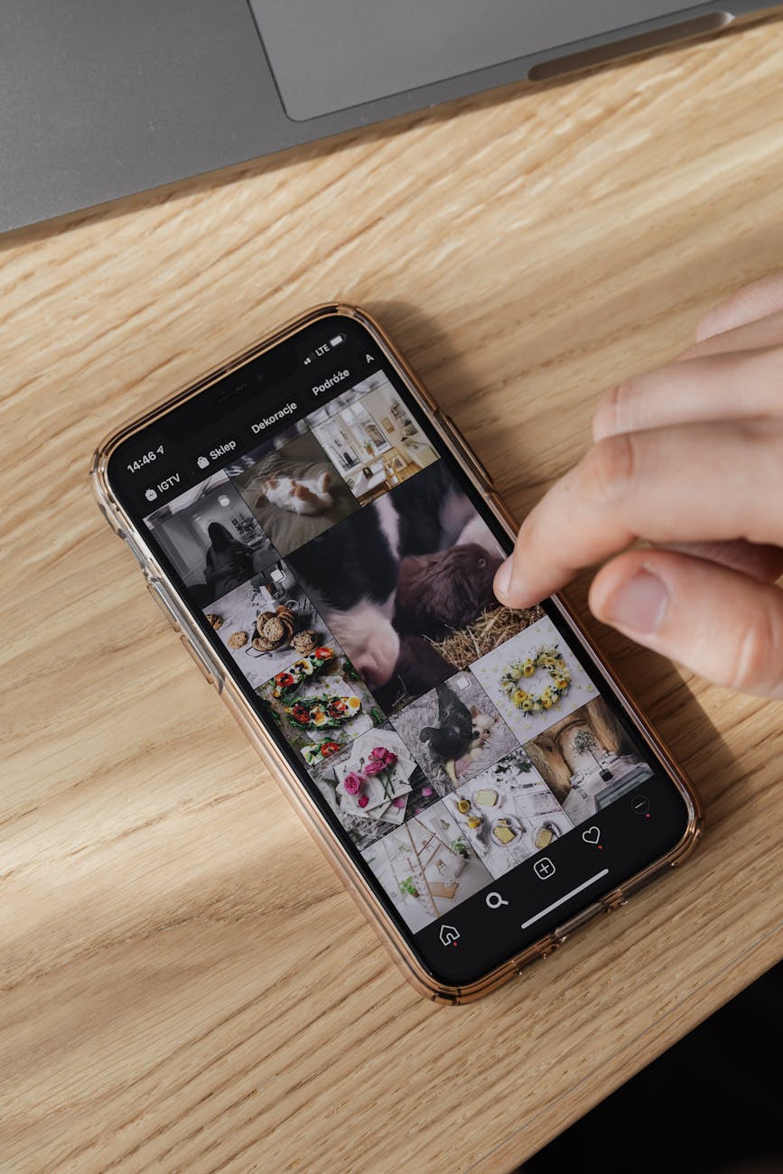 From above of crop unrecognizable person checking pictures in smartphone gallery and scrolling touchscreen cellphone on wooden table near laptop