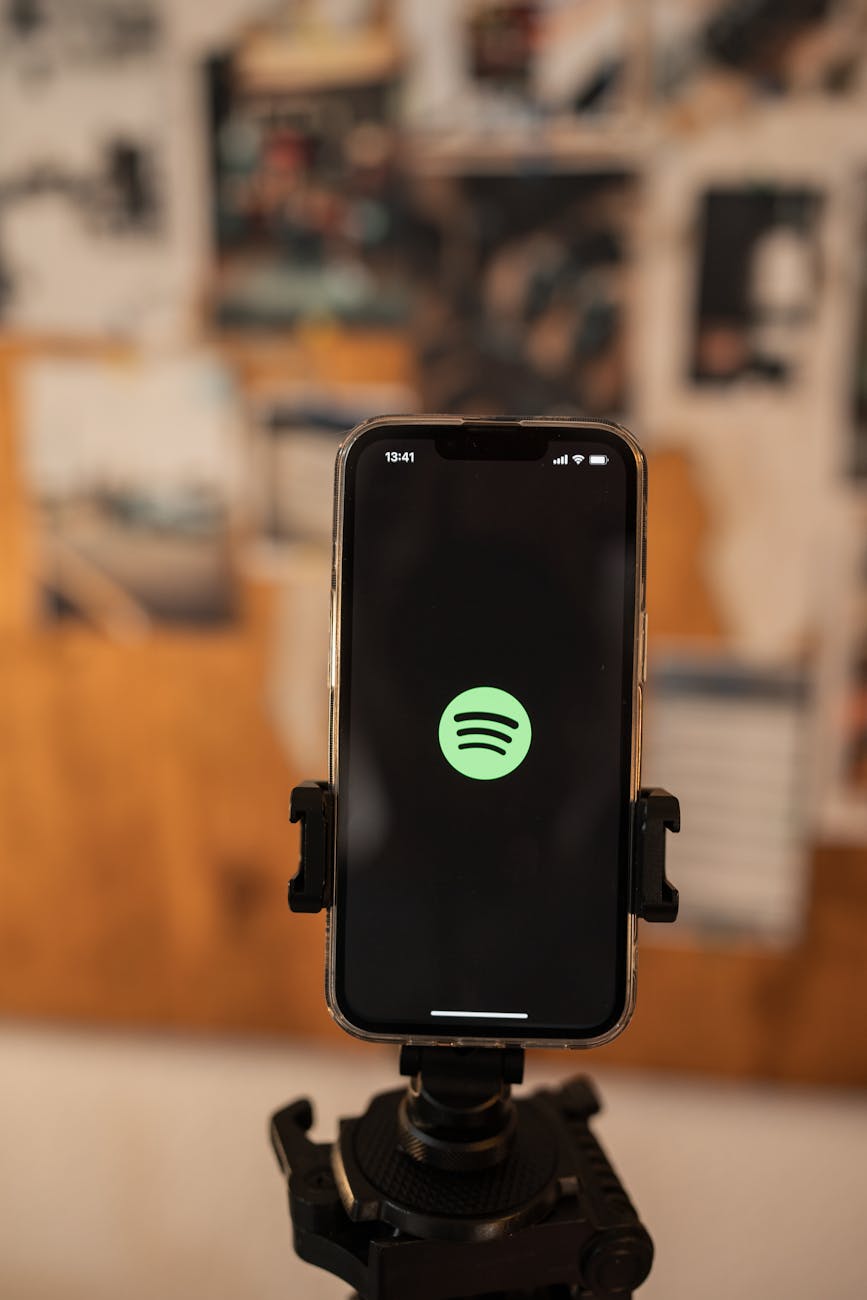 A smartphone showing a music app interface mounted on a stand indoors with blurred photos in the background.