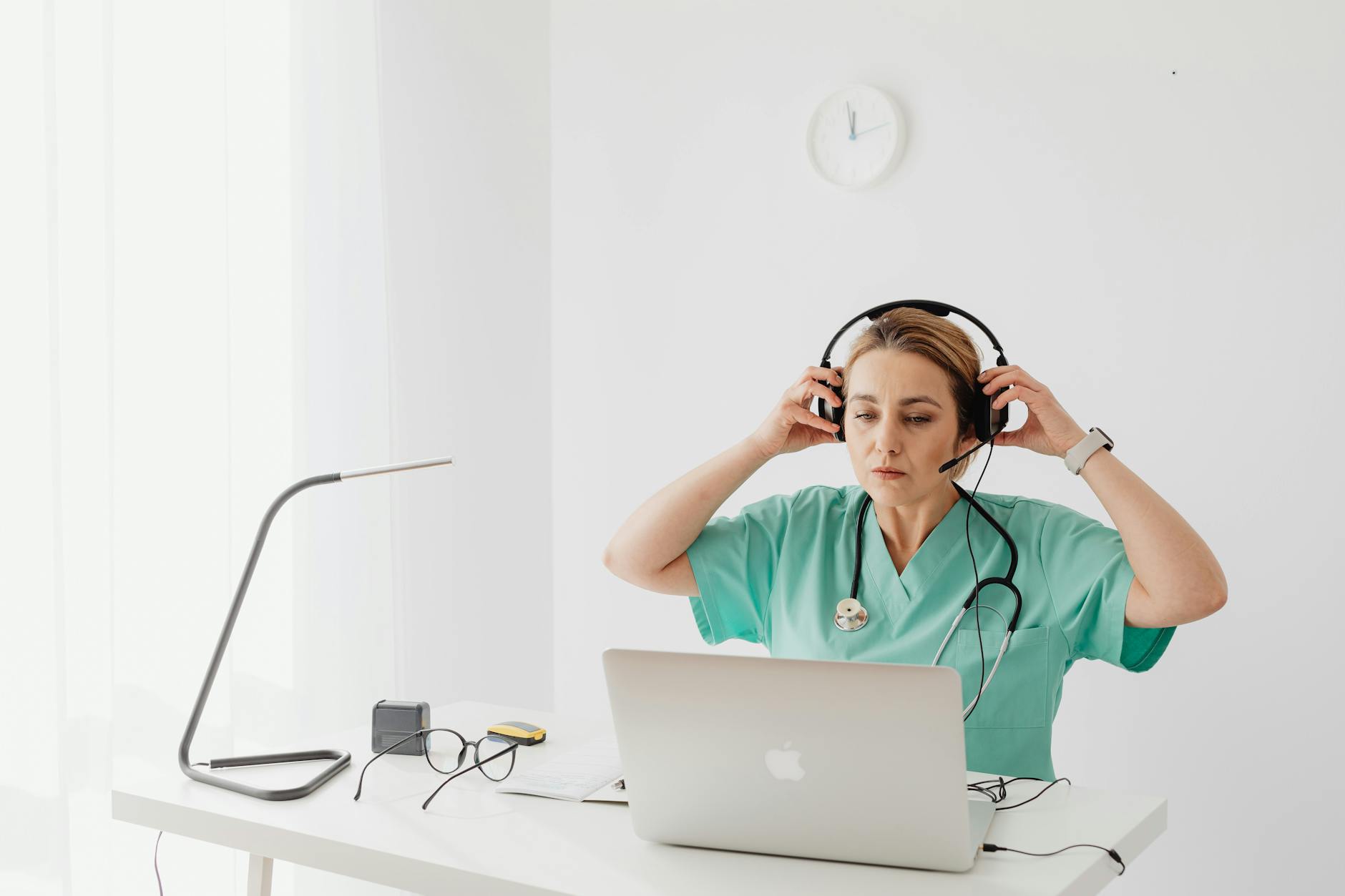 A nurse in teal scrubs using a laptop and headset for remote work at a white desk.
