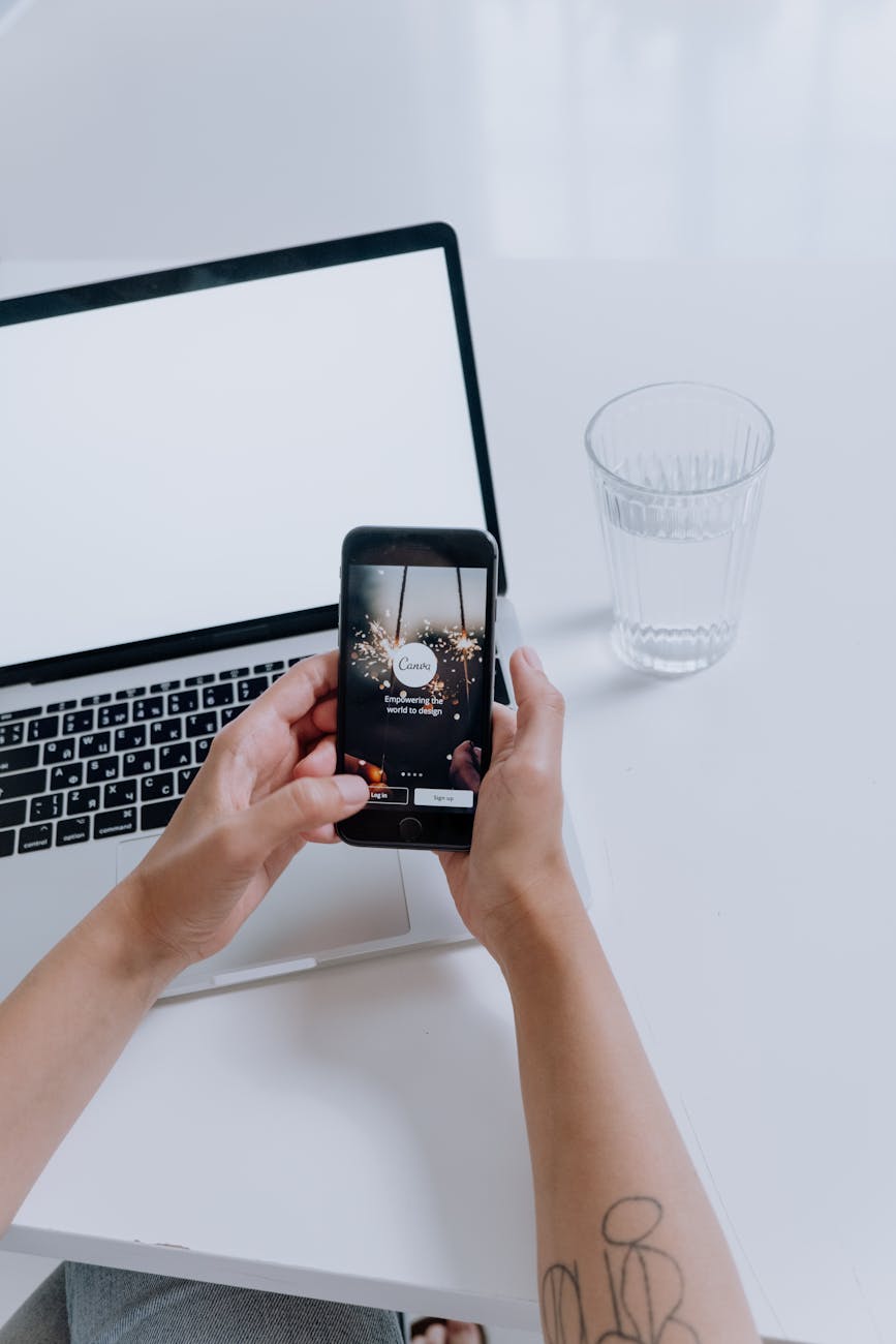 Hands holding a smartphone in front of a laptop. A glass of water is on the table.