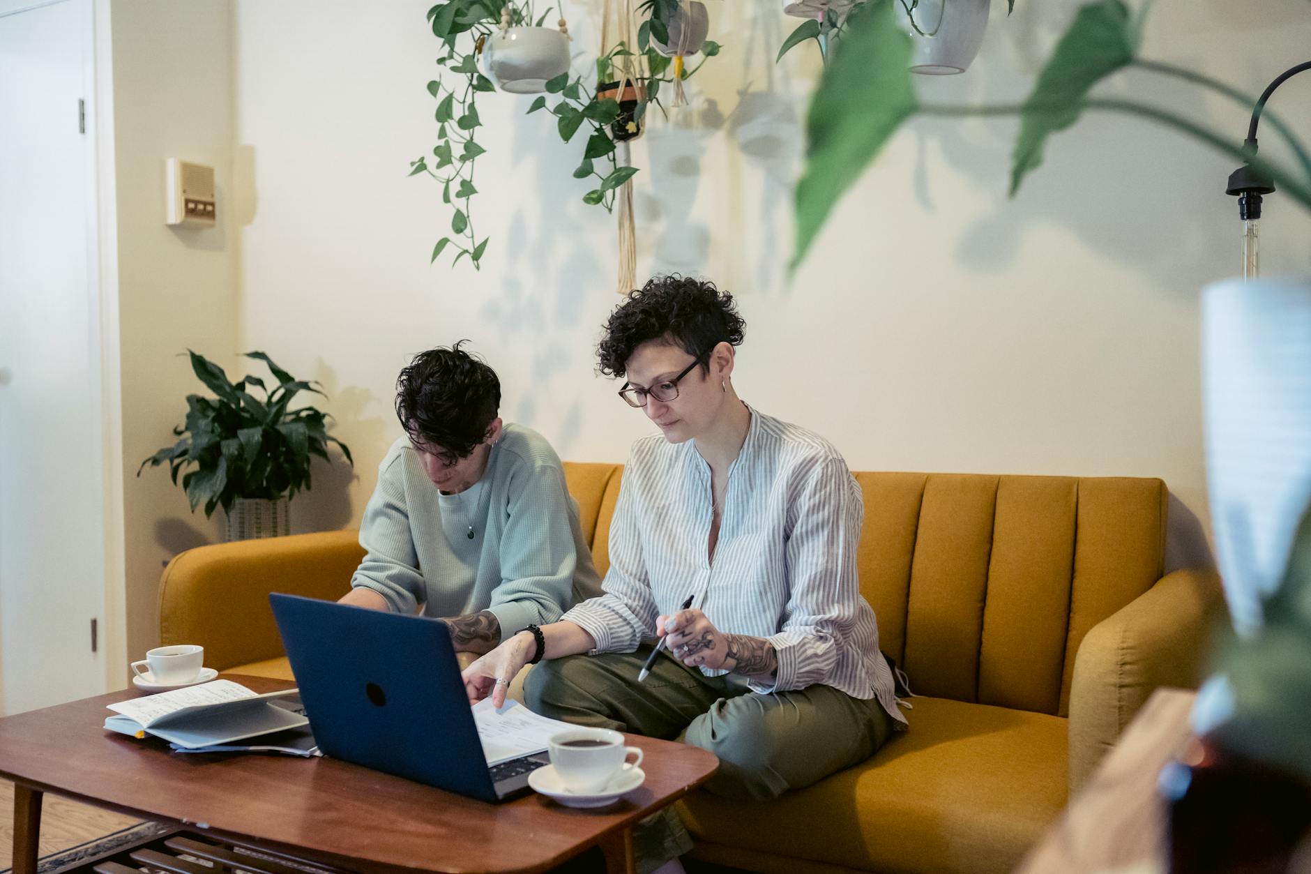 Concentrated young tattooed female freelancers with short dark hair in casual clothes sitting on comfortable sofa and working remotely on laptop