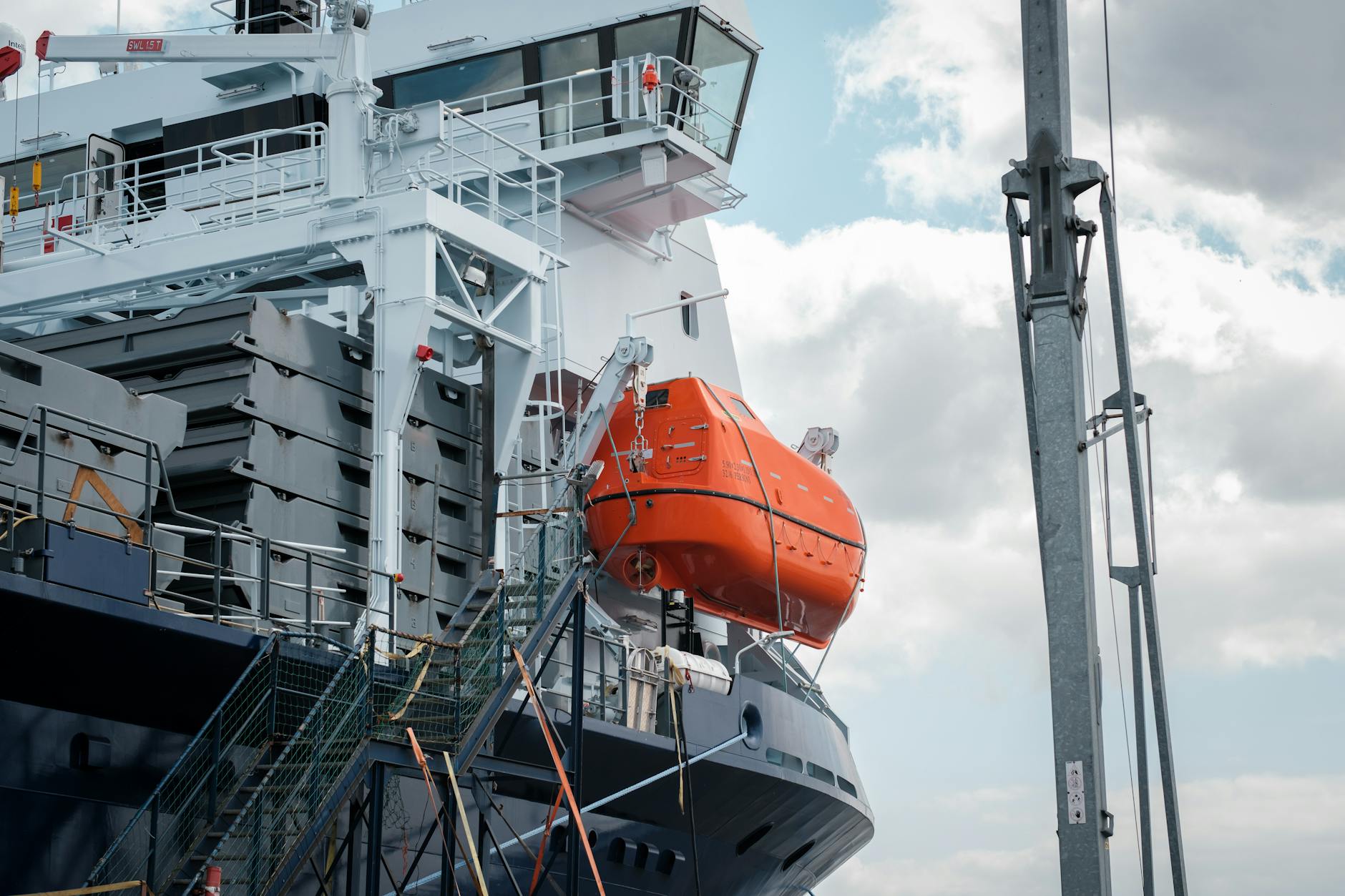 Detailed view of a bright orange lifeboat attached to a cargo ship under a partly cloudy sky.